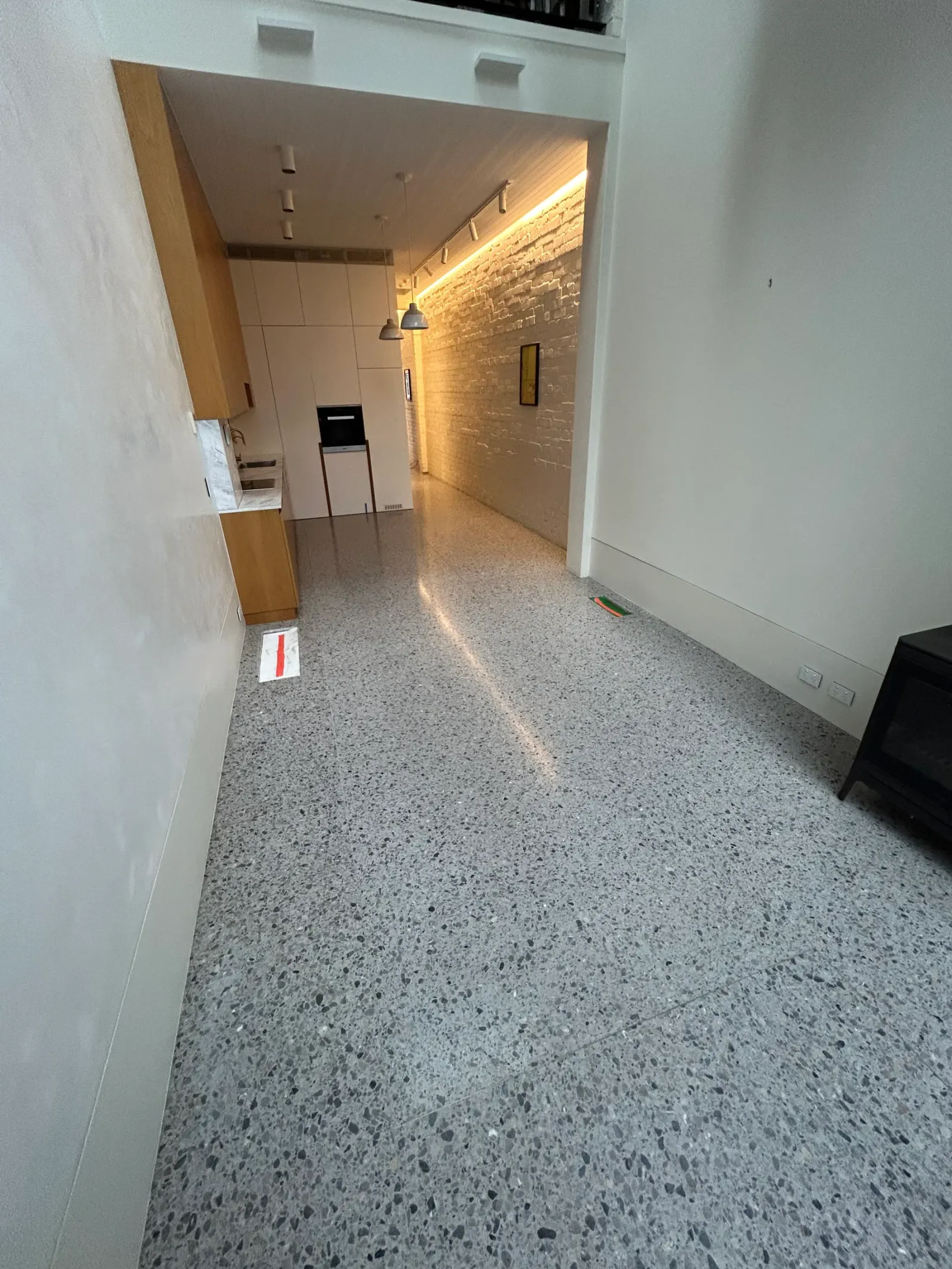 Grey terrazzo floor running from a living space through to a timber kitchen with a backlit brick feature wall