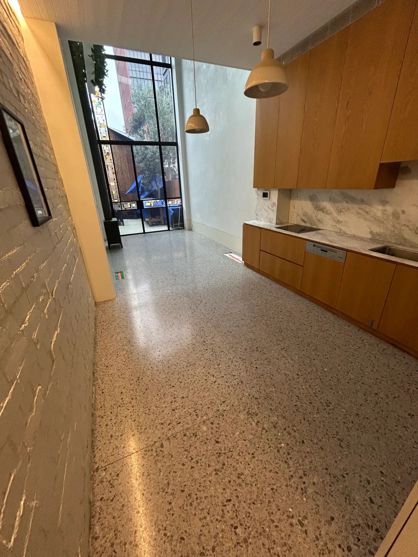 Warm cream terrazzo floor down a galley kitchen with oak joinery, marble splashback and a courtyard window beyond