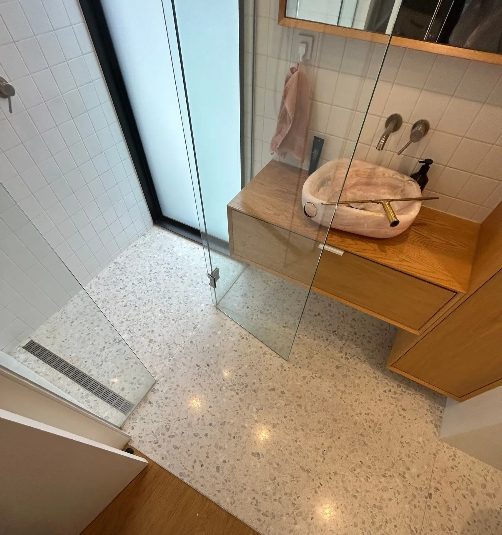 Cream terrazzo bathroom floor running into a walk-in shower with linear drain, oak vanity and stone basin