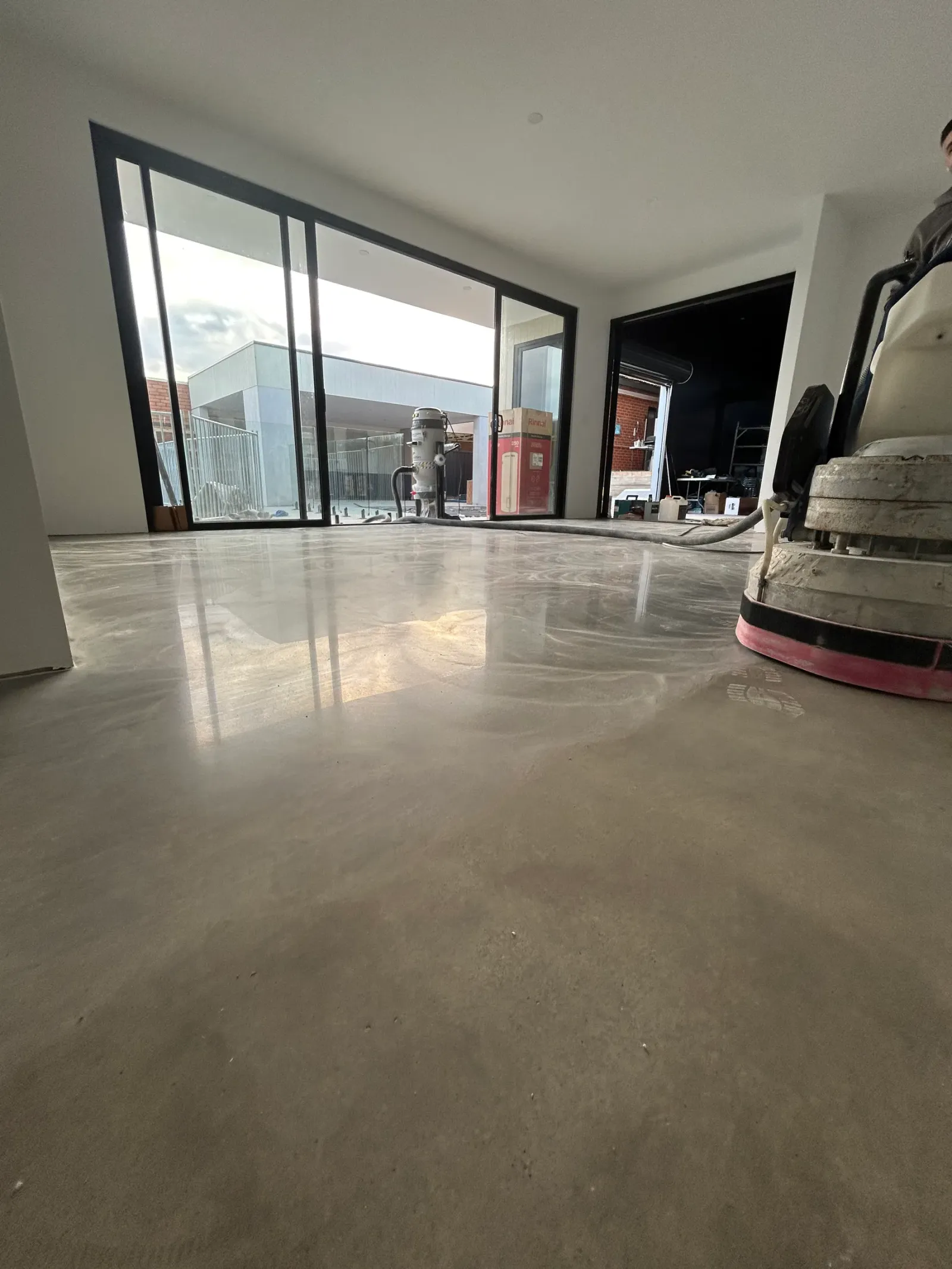 Polished concrete floor mid-job with the grinder head and dust extractor in frame, glazing reflected in the finished surface
