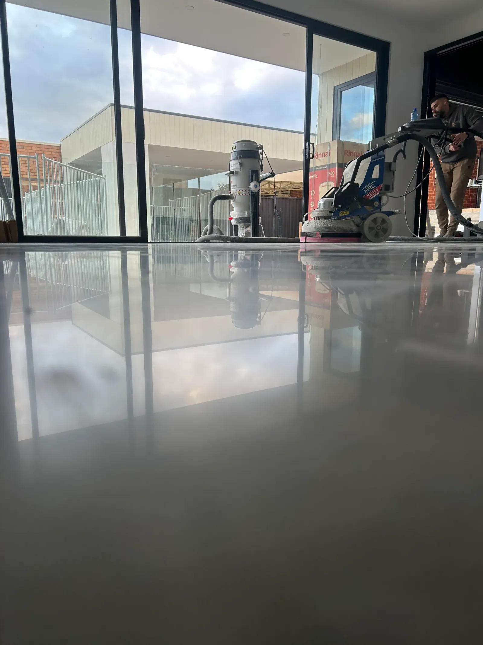 Near-mirror polished concrete floor reflecting the grinder, dust extractor and courtyard glazing in a Melbourne new build