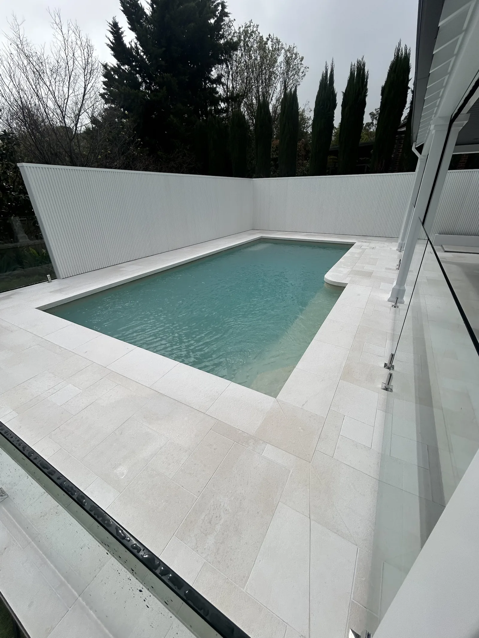 Pale stone paving around a backyard pool with a white batten fence and frameless glass pool fencing, Melbourne