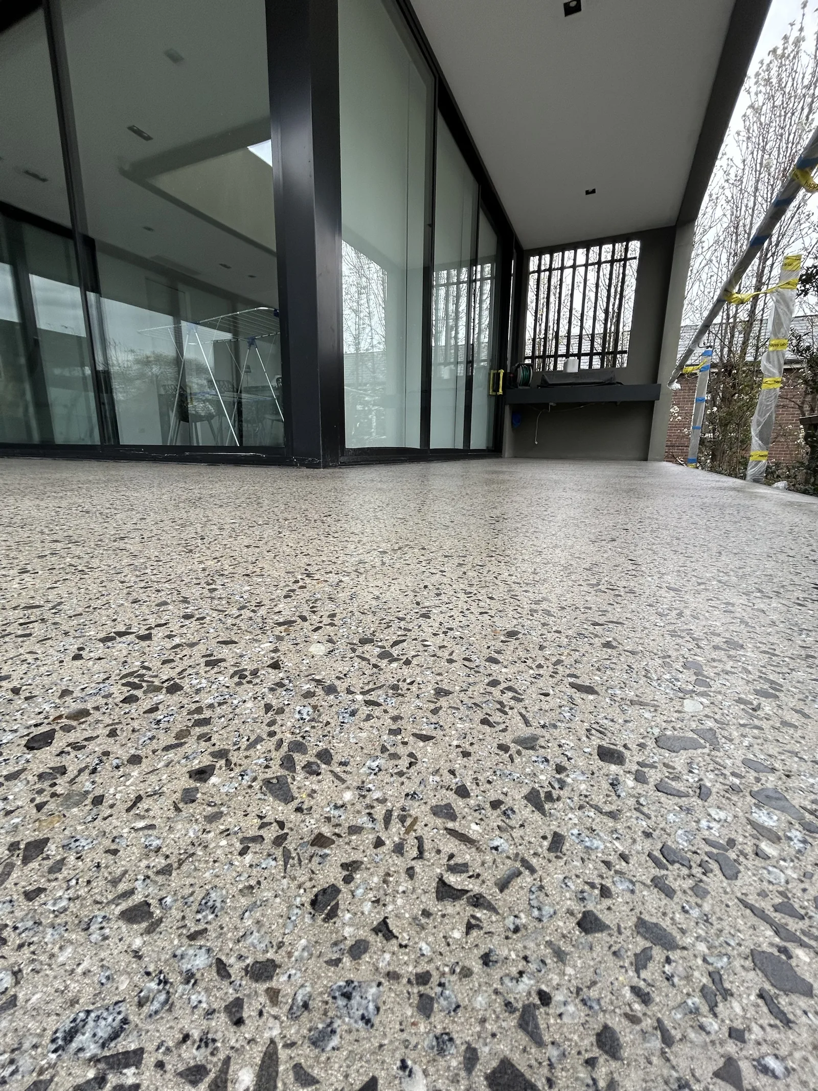Low-angle close-up of concrete with exposed dark and speckled stone aggregate outside a glass-walled home