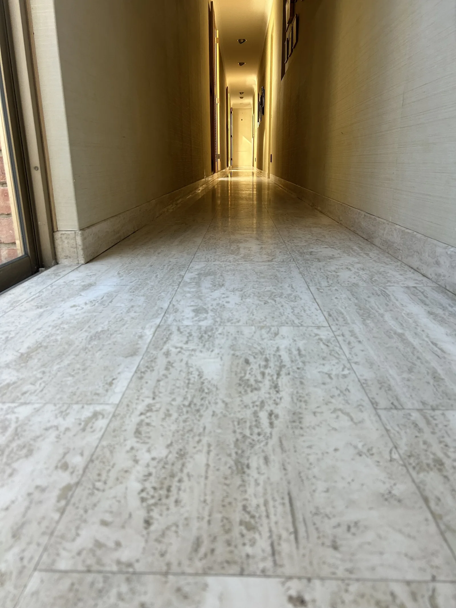 Low-angle view down a long travertine-tiled hallway with a soft sheen and matching travertine skirting