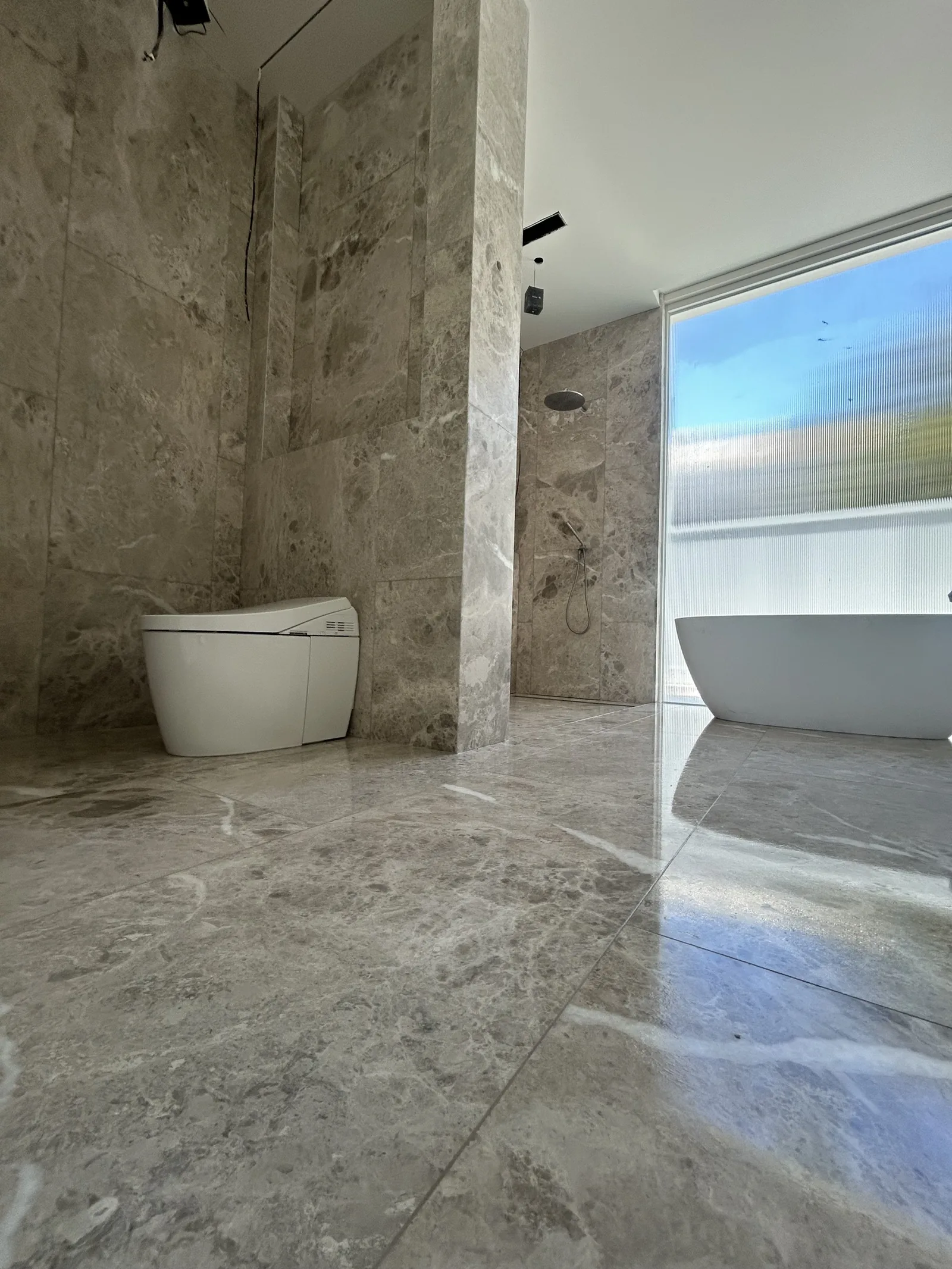 Polished natural stone bathroom floor and walls, a freestanding bath reflected in the floor beside a tall window